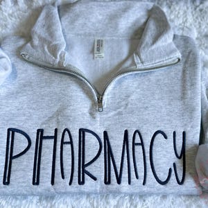 May include: Light gray quarter-zip sweatshirt with the word "PHARMACY" embroidered in navy blue. The sweatshirt has a high collar and a silver zipper. The sleeves are cuffed. The sweatshirt is made of a soft material.