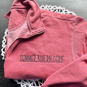 May include: A dusty rose quarter-zip sweatshirt featuring a high collar and a metal zipper. The garment is embroidered with the black text "SUMMER, BSN, RN, CCRN" on the left chest area. The fabric has a soft, worn-in texture and long sleeves.