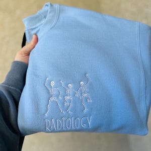 Może przedstawiać: Jasno błękitna bluza z trzema białymi haftowanymi szkieletami tańczącymi i napisem "RADIOLOGY" pod spodem.