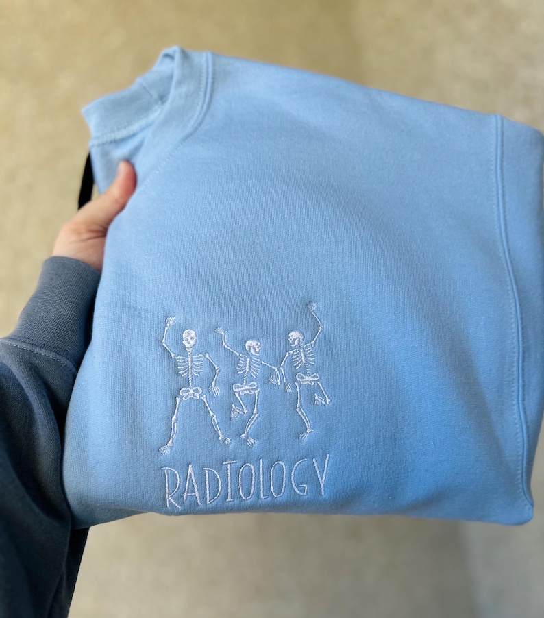 Może przedstawiać: Jasno błękitna bluza z trzema białymi haftowanymi szkieletami tańczącymi i napisem "RADIOLOGY" pod spodem.
