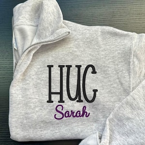 May include: A light grey quarter-zip sweatshirt featuring the word "HUG" in black block letters and the name "Sarah" embroidered in purple script. The sweatshirt has a high collar and a silver zip.