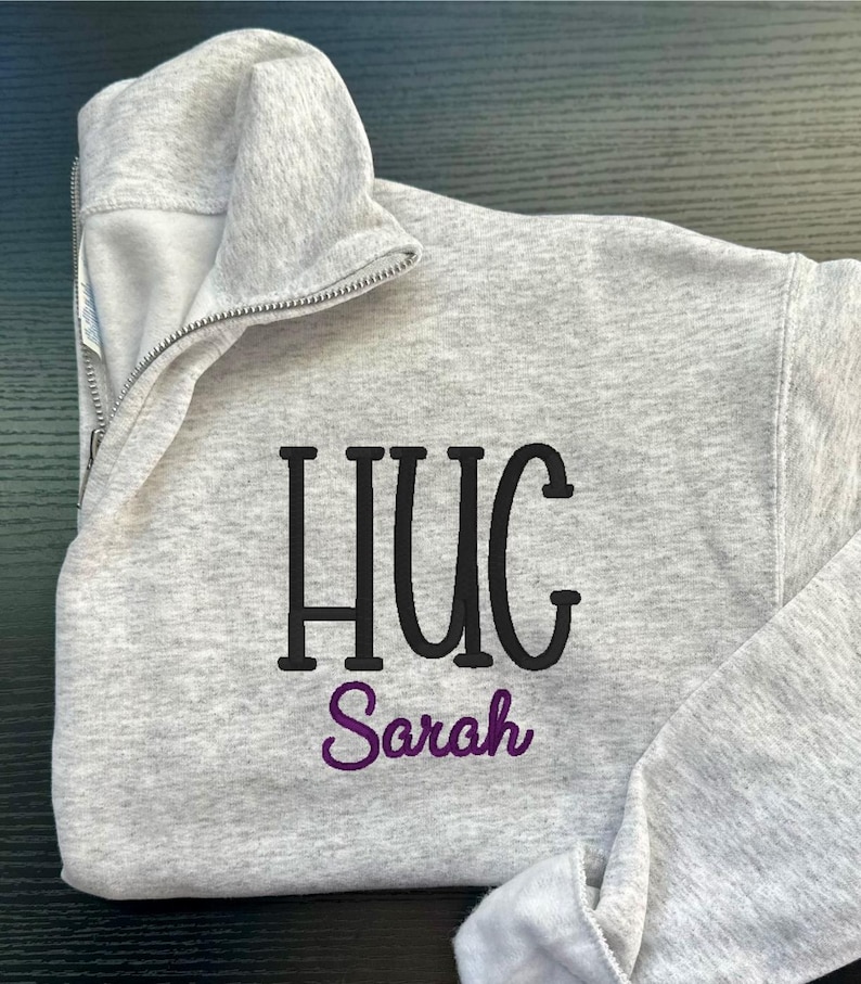 May include: A light grey quarter-zip sweatshirt featuring the word "HUG" in black block letters and the name "Sarah" embroidered in purple script. The sweatshirt has a high collar and a silver zip.