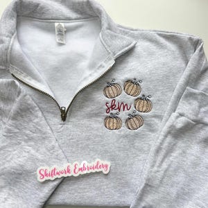 May include: A light gray quarter-zip sweatshirt with embroidered pumpkins and the initials "skm" in burgundy thread. The sweatshirt has a zipper and a tag. The sticker on the bottom says "Shiftwork Embroidery".