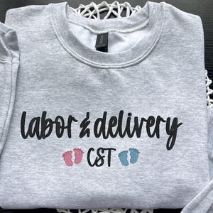 May include: A light gray sweatshirt with the words "labor & delivery" in black script. Below the text are the letters "CST" and two pairs of baby footprints, one pink and one blue.