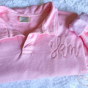 May include: A light pink polo shirt with a collared neckline and a three-button placket. The shirt features embroidered initials "skm" in a cursive font. The shirt is folded on a white, textured surface.