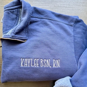 May include: A periwinkle blue quarter-zip sweatshirt with the embroidered text "KAYLEE BSN, RN" in white. The sweatshirt has a gold-colored zipper and a soft, textured appearance. The collar is folded down, revealing a lighter blue lining.