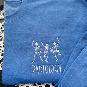 Może przedstawiać: Niebieski t-shirt z długim rękawem z białym haftem przedstawiającym trzy tańczące szkielety i napisem "RADIOLOGY" poniżej.