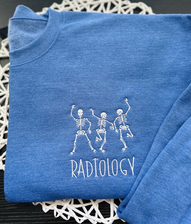 Może przedstawiać: Niebieski t-shirt z długim rękawem z białym haftem przedstawiającym trzy tańczące szkielety i napisem "RADIOLOGY" poniżej.