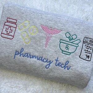May include: A gray sweatshirt embroidered with pharmacy-related symbols: a pill bottle, pills, a caduceus, a mortar and pestle, and a prescription. The words "pharmacy tech" are embroidered in blue script.