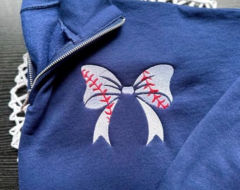 May include: A navy blue sweatshirt with a white baseball-themed bow embroidered on the front. The bow has red stitching to represent the baseball seams.