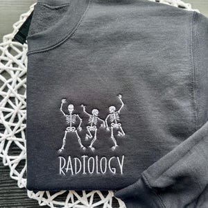 Może przedstawiać: Czarna bluza z białym haftem przedstawiającym trzy tańczące szkielety i napisem "RADIOLOGY" pod spodem.