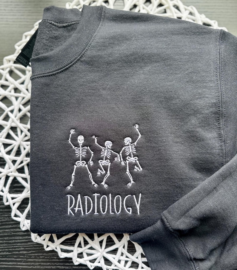 Może przedstawiać: Czarna bluza z białym haftem przedstawiającym trzy tańczące szkielety i napisem "RADIOLOGY" pod spodem.