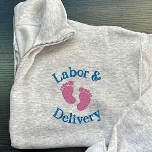 K&ouml;nnte beinhalten: Grauer Sweatshirt mit einem blauen gestickten Design, das "Arbeit & Entbindung" mit rosa Baby-Fu&szlig;abdr&uuml;cken sagt.