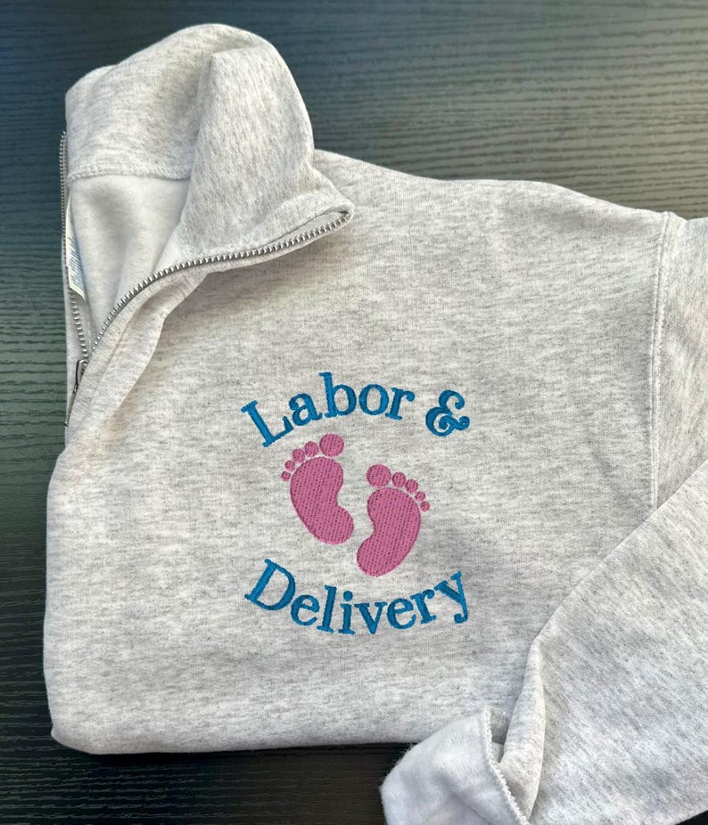 K&ouml;nnte beinhalten: Grauer Sweatshirt mit einem blauen gestickten Design, das "Arbeit & Entbindung" mit rosa Baby-Fu&szlig;abdr&uuml;cken sagt.