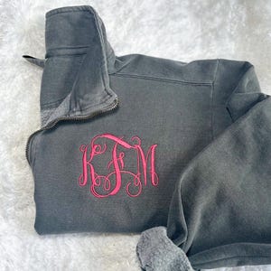 May include: A gray pullover sweatshirt with a quarter-zip neckline and a pink embroidered monogram. The sweatshirt is made of a soft material and has a relaxed fit. The monogram features the letters KFM.