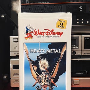 May include: A VHS tape case with a white background and a colorful image of a woman riding a winged creature. The case has the title "Heavy Metal" and the Walt Disney Home Video logo. The case is rated G for general audiences.