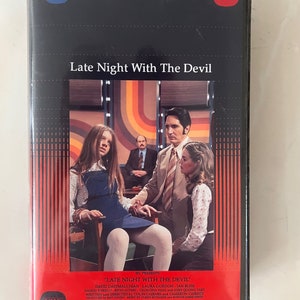 May include: VHS video cassette cover for the horror film "Late Night With The Devil". The cover features a scene from the film with three people in a room with a retro 70s design. The text "IFC Presents" is at the bottom of the cover.