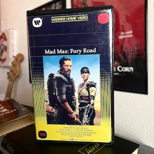 May include: A VHS tape case for the movie "Mad Max: Fury Road" featuring Tom Hardy and Charlize Theron. The case is black with a red "Action" label and a Warner Bros. Home Video logo.