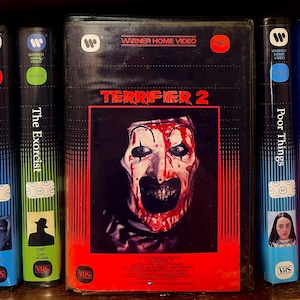 May include: A VHS tape of the horror film "Terrifier 2" featuring a close-up of a scary clown face with blood. The tape is in a black case with red accents and the Warner Home Video logo. Other VHS tapes are visible.