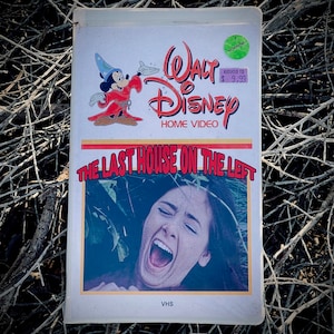 May include: A VHS tape case with a white background and a red and white Walt Disney Home Video logo. The case features a scene from the horror film "The Last House on the Left" with a woman screaming. The case has a sticker that says "Horror" and "Reduced to $9.99".