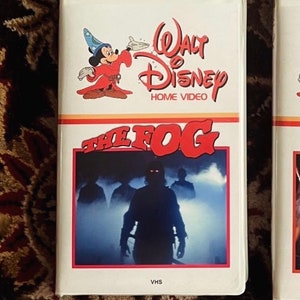May include: A white VHS tape case with a red and white title card that reads "The Fog". The case features a Disney Home Video logo with Mickey Mouse dressed as a wizard. The front of the case shows a scene from the movie with a figure in the fog.