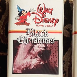 May include: A white VHS tape with a red and black title card that reads "Walt Disney Home Video Black Christmas". The cover features a cartoon image of Mickey Mouse wearing a red robe and a blue hat. The bottom of the cover shows a scene from the movie with a woman in a white dress with a plastic bag over her head.