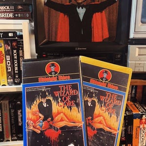 May include: Two VHS tapes of "The Wizard of Gore" are held up in front of a vintage television. The tapes have a black and yellow design with the film title and a graphic of a magician. The TV shows a man in a tuxedo and cape.