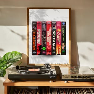 May include: A framed print displaying a selection of classic horror film VHS tapes, such as "Halloween," "Psycho," and "Scream." The tapes are aligned vertically, showcasing their titles. A record player and stereo equipment are in the foreground.