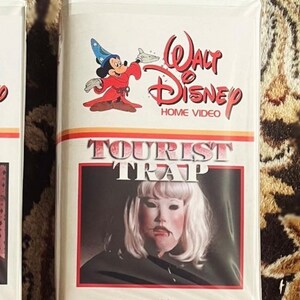 May include: A VHS tape with a white background and a red, white, and blue Walt Disney Home Video logo. The title of the movie is "Tourist Trap" in red and black letters. The cover features a woman with white hair and a white mask, wearing a black cloak. The text "VHS" is printed in black at the bottom of the cover.