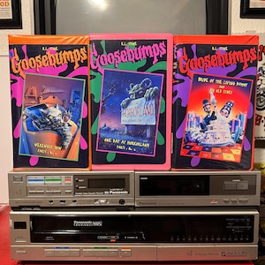 May include: Three Goosebumps VHS tapes with colorful covers featuring illustrations of a werewolf, a monster, and a bride and groom. The tapes are stacked on top of two vintage Panasonic VHS players.