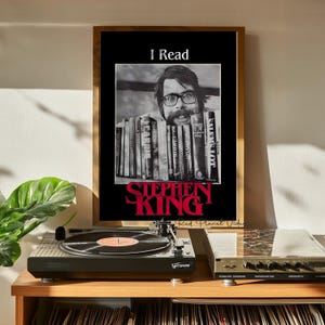 May include: A black and white poster with a framed border. The poster features a photo of Stephen King with a stack of books in front of his face. The text "I Read" is at the top of the poster and "Stephen King" is at the bottom in red letters.