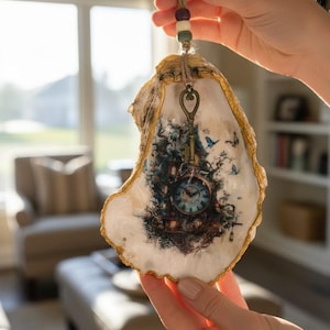 May include: A decorative oyster shell ornament with gold-painted edges, featuring a printed image of a clock, trees, and blue butterflies. A vintage-style metal key hangs from the top, suspended by a jute twine cord with small wooden beads.