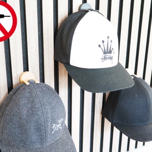 Könnte beinhalten: Drei Baseballkappen hängen an einem an der Wand montierten Hutständer. Die Kappen sind schwarz, grau und schwarz-weiß mit einem Kronenlogo. Der Ständer ist aus Holz mit schwarzen vertikalen Latten.