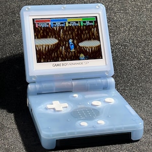 Puede incluir: Una consola de juegos portátil Game Boy Advance SP azul claro, abierta y mostrando un videojuego colorido. La consola presenta un borde de pantalla blanco, botones blancos y el texto "GAME BOY ADVANCE SP".