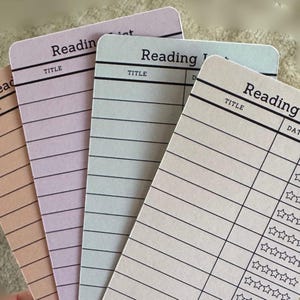 May include: Four pastel-colored reading list cards with rounded corners. Each card has the word "Reading" at the top, with lines for "Title" and "Date". One card has a rating section with stars. The cards are in shades of peach, lavender, and mint.