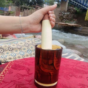 Peut inclure: Un ensemble de mortier et pilon en bois. Le mortier est un cylindre brun foncé avec une base et un intérieur de couleur claire. Le pilon est de couleur claire avec un manche brun foncé. L'ensemble est sur une nappe rouge.