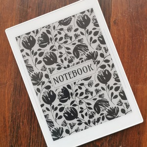 以下が含まれることがあります： 画面に白黒の花柄が描かれた白い電子書籍リーダー。画面には「NOTEBOOK」という文字が表示されています。
