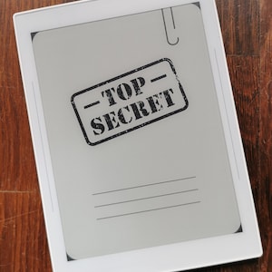以下が含まれることがあります： 白い電子書籍リーダーに、黒い「TOP SECRET」のスタンプが画面に押されています。電子書籍リーダーは白いベゼルと黒い画面を持っています。