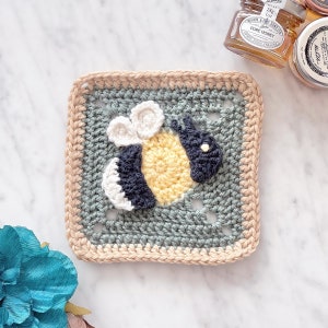 May include: A square, crocheted coaster featuring a bee design. The bee has a yellow body, white wings, and a navy blue head and stripes. The coaster has a light blue center, a tan border, and a cream-colored edge.