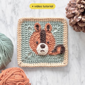 May include: A square, crocheted piece featuring a brown and cream-colored bear face on a light blue background. The bear has a cream-colored snout and a dark brown stripe down its face. The border is a light tan color. The text "+ video tutorial" is in the upper left corner.