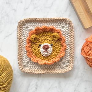 May include: A crocheted square with a lion face in the center. The lion is made with yellow yarn and has a brown nose and black eyes. The square is made with beige and white yarn.