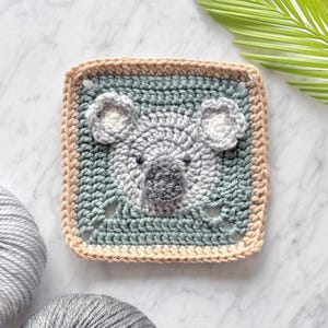 May include: A crocheted square featuring a koala face in shades of gray and green. The koala has a light gray face with a dark gray nose and ears. The square is bordered with a light brown crocheted edge.