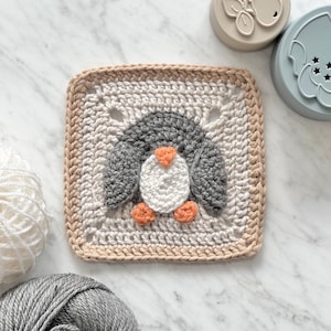 Puede incluir: Un cuadrado tejido a crochet con un diseño de pingüino. El pingüino está hecho con hilo gris, blanco y naranja. El cuadrado está rodeado por un borde beige.
