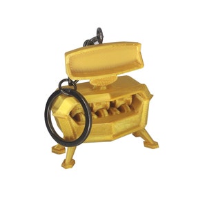 Può includere: Un portachiavi robot stampato in 3D color oro con un anello in metallo nero. Il robot ha una testa rettangolare con una parte superiore piatta e un corpo con una serie di ingranaggi visibili sul davanti.