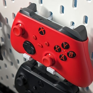 May include: A red Xbox controller with black buttons and a white mounting bracket. The controller is mounted on a white pegboard wall.