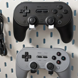 May include: Two 8BitDo Pro 2 controllers, one black and one grey, are mounted on a pegboard wall. Both controllers have a D-pad, four face buttons, two shoulder buttons, and two analogue sticks.