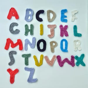 May include: A selection of colourful, crocheted alphabet letters displayed on a white background. The letters are in a variety of shades, including red, blue, green, yellow, and pink. The letters have a soft, textured appearance, creating a playful and decorative effect.