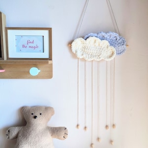 May include: A white and gray crocheted cloud mobile with wooden beads hanging from it. The mobile is hanging from a white wall. The text "find the magic" is written on a framed picture on the wall.
