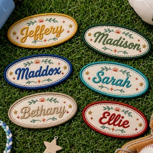 May include: Six oval-shaped embroidered patches featuring off-white fabric with personalized names in various thread colors including yellow, green, blue, teal, tan, and red, accented with small embroidered stars, leaves, and sun motifs.