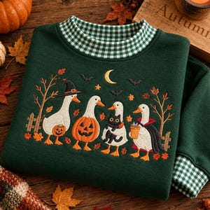 May include: A dark green sweatshirt featuring an embroidered scene of four white geese in costumes, including a witch hat, a pumpkin suit, a black cat, and a vampire cape. The collar and cuffs have a green and white gingham pattern.